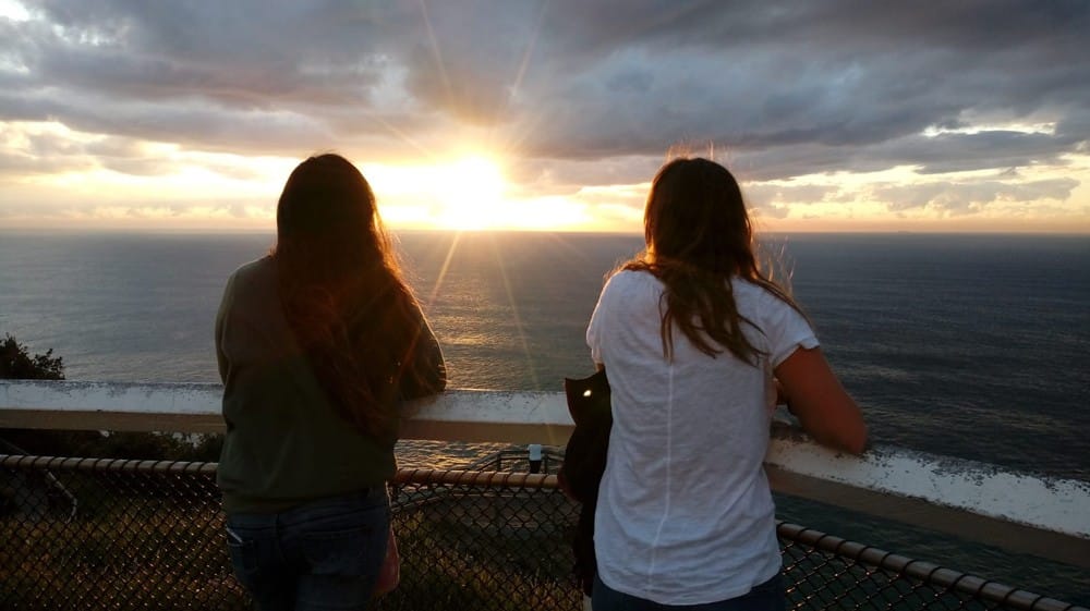 dos chicas morenas y de espaldas apoyadas en una barandilla de madera mirando la puesta de sol encima del mar